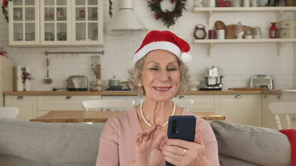 A positive elderly woman in a Christmas hat communicates online with her relatives via a video call on her smartphone, congratulates them on the holidays and sends greetings.