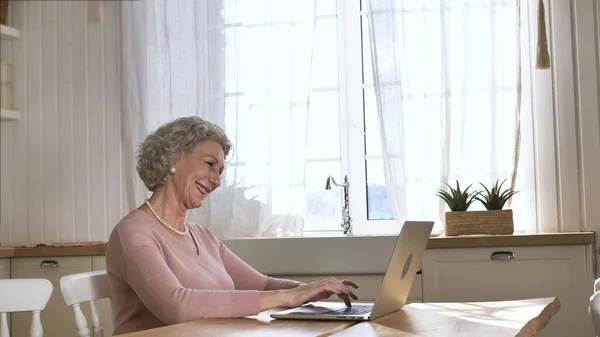 Стара жінка радіє підняттю рук, сидячи біля ноутбука на кухні — стокове фото