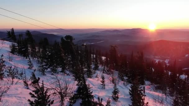 눈덮인 나무 꼭대기를 지나 하늘을 떠다니며 일몰에는 산을 배경으로 한 스키 리프트 객실을 넘어 떨어지는 태양을 배경으로 날아간다. — 비디오