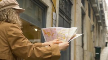 Kahverengi ceketli turist elinde haritayla dar Avrupa caddelerinde yürüyor