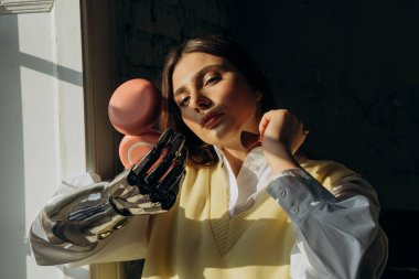 Biyonik protez kollu çekici bir kadın pencerede duran bir fırçayla makyaj yapar ve küçük bir aynaya bakar.