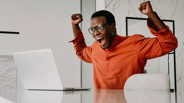 Happy african american man freelancer rejoices working at laptop from home, joyful black man with glasses won the jackpot raised his hands up looking at the computer