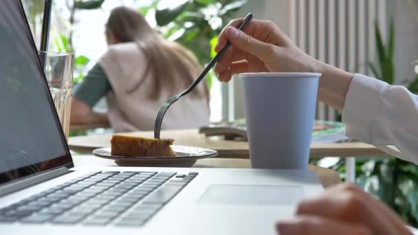 Kvinna med kaffe och laptop tar bit av kakan vid bordet — Stockvideo