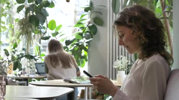 Glad ung kvinna sms på telefon sitter i elegant café — Stockvideo