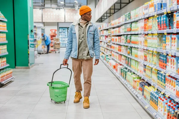 Афроамериканец ходит с пустой зеленой корзиной через супермаркет и выбирает продукты — стоковое фото