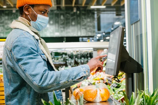 Afrikkalainen mies lääketieteellisessä naamiossa painaa tuoreita appelsiineja sähköisessä mittakaavassa supermarketissa. — kuvapankkivalokuva