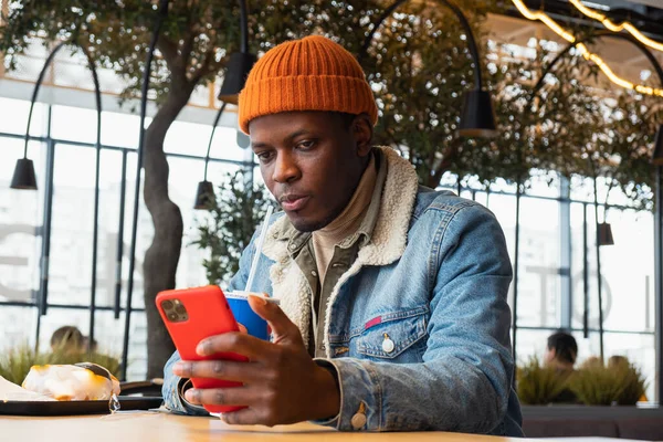 Black man in knitted hat and jacket uses smartphone in cafe