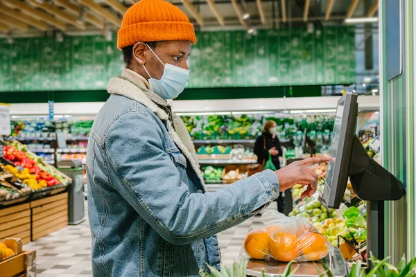Afroamerikansk fyr i maske vejer appelsiner i supermarkedet - Stock-foto