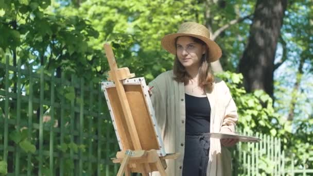 Jonge vrouw kunstenaar in bruine hoed staat bij ezel en verf — Stockvideo