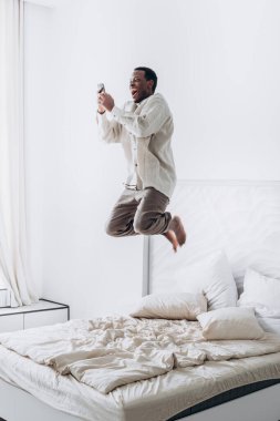 Komik Afro-Amerikalı adam büyük bir yatakta telefonla zıplıyor.