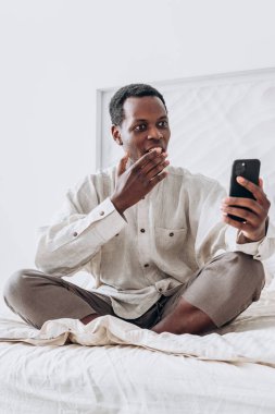 Gülümseyen siyah adam video çağrısında el sallıyor, yatakta oturuyor.