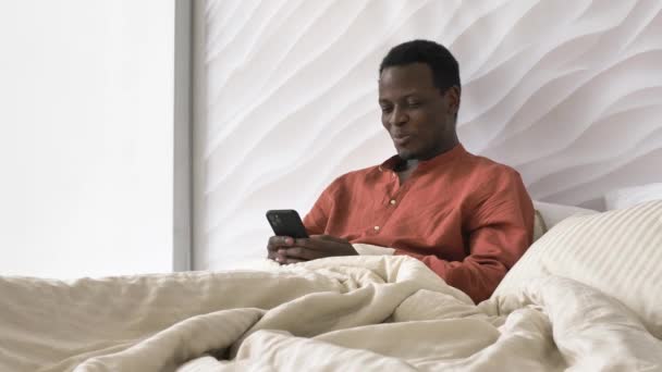 Афроамериканец смеется, читая телефонные сообщения в постели — стоковое видео