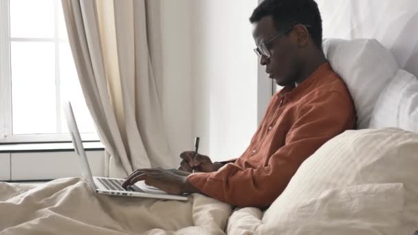 Homem afro-americano com óculos faz anotações trabalhando no laptop — Vídeo de Stock