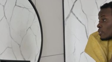 Afro-Amerikalı adam banyoda aynanın yanında yüzünü siliyor.