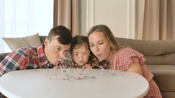 Мать отец и маленькая девочка дуют конфетти в гостиной — стоковое видео