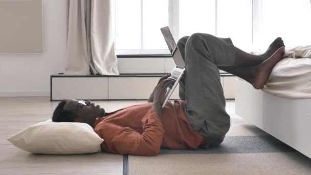 Лінивий афроамериканець працює на ноутбуці, лежачи на підлозі. — стокове відео