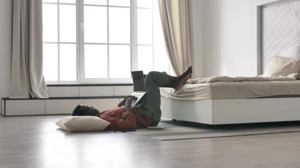 El freelancer afroamericano trabaja en casa en un portátil tumbado en el suelo frente a una gran ventana luminosa — Vídeos de Stock