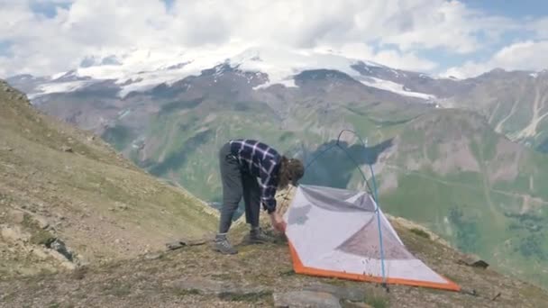 한 여성 여행자가 구름낀 여름날 눈덮인 높은 산의 배경에 텐트를 치고 있다 — 비디오