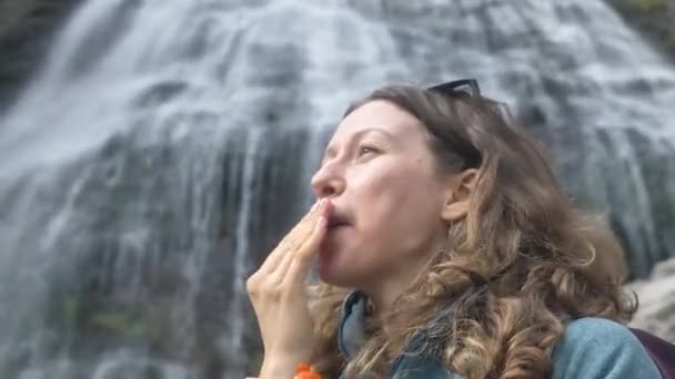활동적 인 한 여성 관광객 이 산 속에 있는 큰 폭포의 뒤 편에서 일광욕으로 얼굴을 때리고 있다 — 비디오