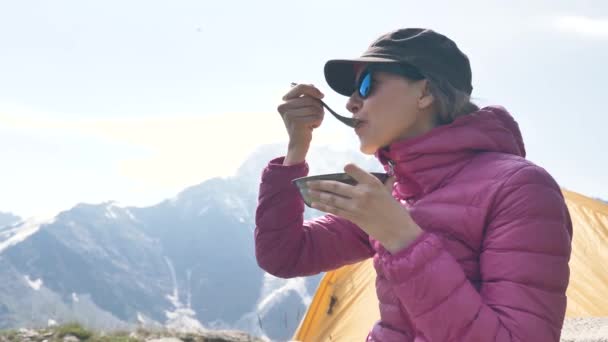 Молода жінка, яка подорожує в рожевій куртці, снідає з кашею в горах біля намету — стокове відео