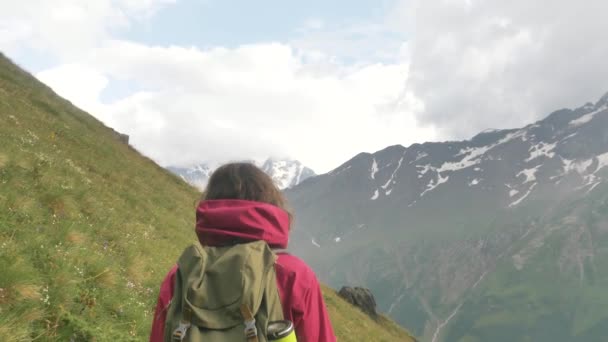 Uma turista caminha ao longo de uma encosta verde em um caminho de montanha contra o fundo de picos cobertos de neve com uma mochila — Vídeo de Stock