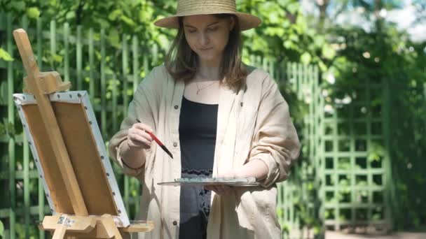 Joven artista en sombrero marrón se encuentra en caballete y pinturas — Vídeos de Stock