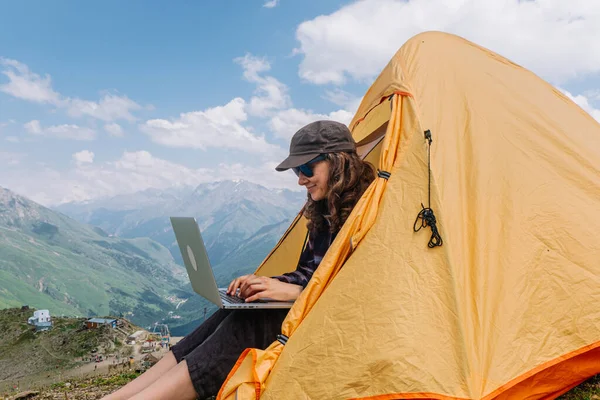 Uma mulher freelance feliz e livre está digitando em um laptop enquanto está sentado em uma tenda em um acampamento alto nas montanhas — Fotografia de Stock