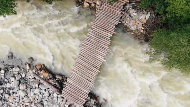 Uma viajante feminina com uma mochila caminha em uma ponte de madeira através de uma vista aérea tempestuosa do rio da montanha — Vídeo de Stock