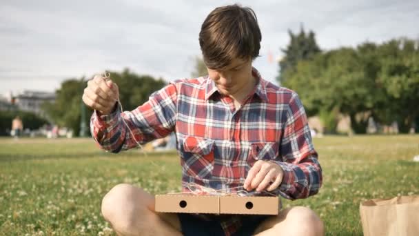 Człowiek otwiera tekturowe pudełko i czuje pizzę siedzącą na trawie — Wideo stockowe
