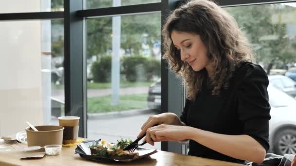 Jonge mooie vrouw met krullend haar dineert in een café, snijdt een zalmbroodje met een tafelmes — Stockvideo