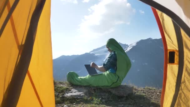 Positieve freelance man in een slaapzak opent een laptop en begint te werken bij dageraad in de bergen, schieten vanuit een tent — Stockvideo