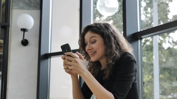 Красива жінка з кучерявим волоссям отримала дивовижне радісне повідомлення на своєму мобільному телефоні і з радістю сидить на тлі великого вікна — стокове відео