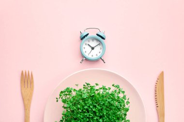 Çalar saat, çatal bıçak takımı ve yeşillikli tabak. Pembe renk. Aralıklı oruç, öğle yemeği ve diyet konsepti.