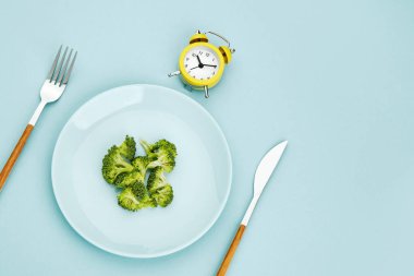 Aralıklı perhiz, diyet, öğle yemeği konsepti. Sarı alarm saati, tabakta brokoli ve mavi arka planda çatal bıçak takımı..