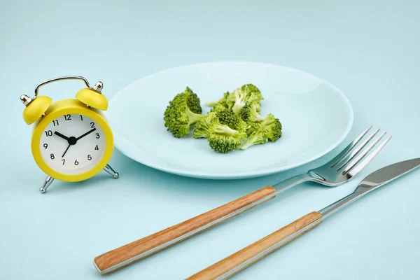 断断续续的禁食 午餐时间概念 黄色闹钟 盘子里有花椰菜 蓝色背景上有餐具 — 图库照片