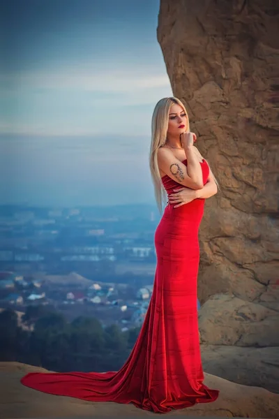 日没の街の背景には リング山の中に長い赤いドレスでブロンドのモデルの女の子 肩には鳥の羽を持つ夢のキャッチャーの入れ墨があります — ストック写真