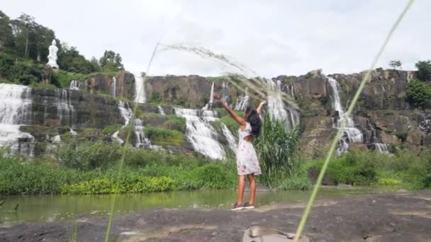 Unbekümmerte Frau im Kleid steht vor einem majestätischen Wasserfall mit viel Grün. — Stockvideo