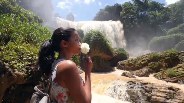 Jonge aziatische vrouw staan in de buurt van een reusachtige waterval ruiken een witte bloem. — Stockvideo