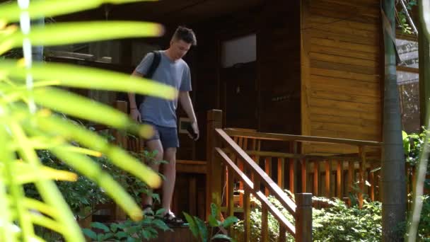 Junger Mann mit Rucksack läuft die Treppe einer Veranda eines rustikalen Hauses hinunter. — Stockvideo