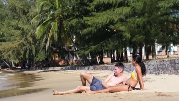Διαφυλετικό έγκυο νεαρό ζευγάρι που βρίσκεται στην άμμο στην παραλία σε μια ηλιόλουστη μέρα. — Αρχείο Βίντεο