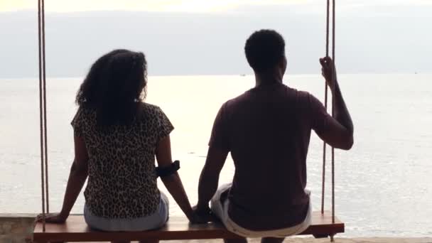 Vista trasera de la joven pareja dulce sentada en un columpio relajándose frente al mar. — Vídeo de stock