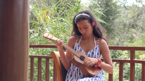 Mujer joven en un vestido sentado en la silla jugando un ukelele al aire libre. — Vídeos de Stock