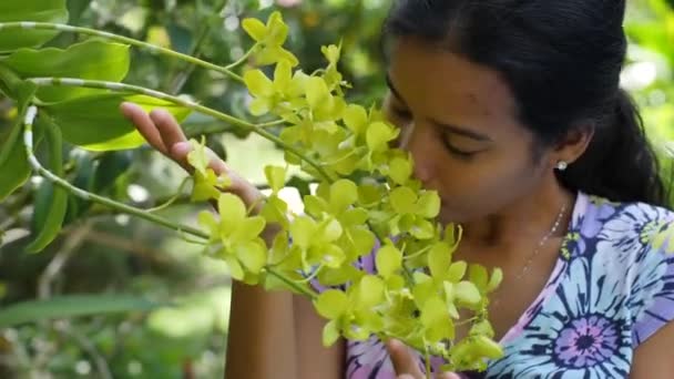 Boldog ázsiai nő érzi a virágok illatát a trópusi kertjében. — Stock videók