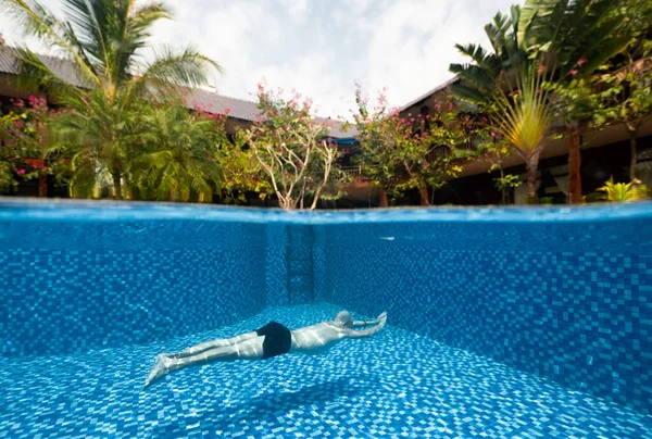 一个男人在游泳池下面游泳，可以看到热带别墅. 免版税图库照片
