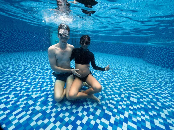 Young pregnant couple in swimwear and sunglasses sitting at the bottom of pool. Royalty Free Stock Photos