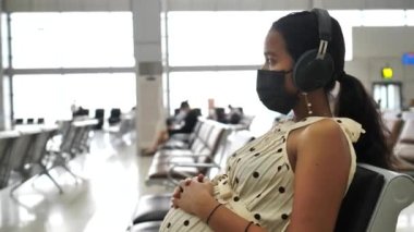 Hamile kadın, maske ve kulaklık takıyor, bekleme salonunda oturuyor..