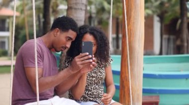 Güzel, genç Afrikalı çift salıncakta oturuyor ve akıllı telefon kullanıyor..