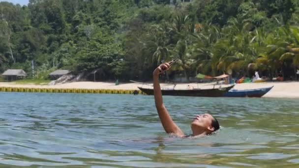 Mujer sosteniendo vídeo de filmación de teléfonos inteligentes mientras yace en el agua en la playa. — Vídeos de Stock