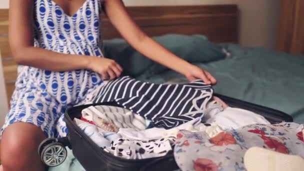 A close up view of a pregnant woman sitting on the bed packing clothes and placing it into the luggage. — Stock Video