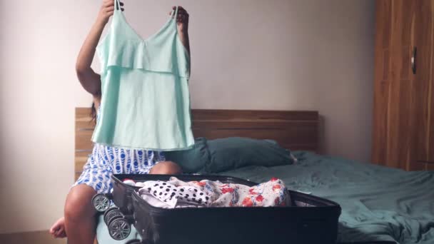 A young pregnant woman sitting on the bed with luggage folding her clothes. — Stock Video